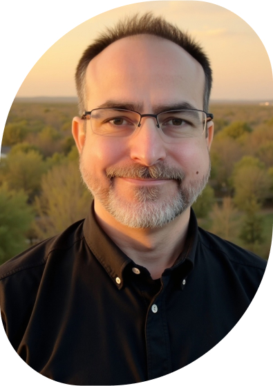 Thomas Sola. White male with a beard and mustache set before a wooded area at sunset. The lighting is turning red. It looks quiet and peaceful.